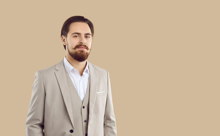 Young confident and stylish man looking at camera with confident look on beige background. Handsome man with mustache and beard dressed in three-piece suit. Concept of confident men and business.の写真素材