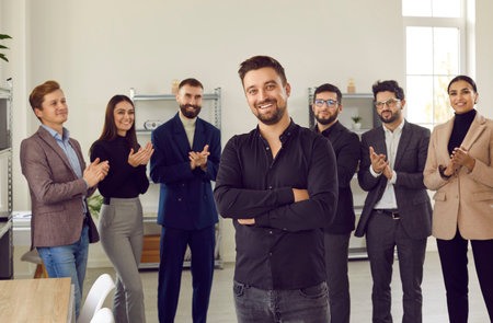Smiling young attractive boss and business leader proudly looking at camera with team of people applauding in background in the office. Business team or group of staff congratulating their leader.の写真素材