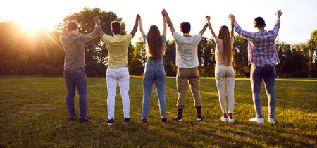 Diverse friends meet in the park. Group of multiracial young people standing in row on green lawn and holding hands up in evening sunlight. Back view, from behind. Friendship, community conceptの写真素材