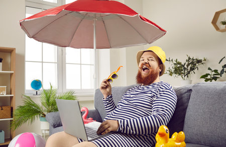 Funny happy bearded young man in sun hat sitting on couch with beach umbrella and inflatable toys, using laptop, browsing best cheap prices online and booking tickets for summer holiday travelの写真素材