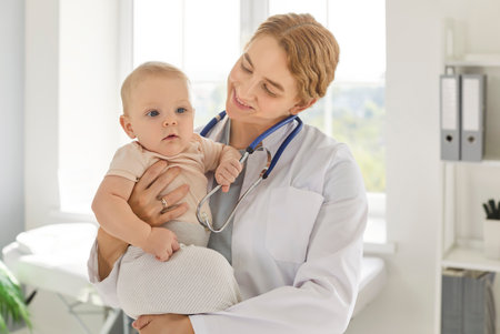 Young smiling female pediatrician holding toddler baby on her hands during medical checkup in clinic. Professional caring doctor creating warm and comforting atmosphere for newborn child. Baby health.の写真素材