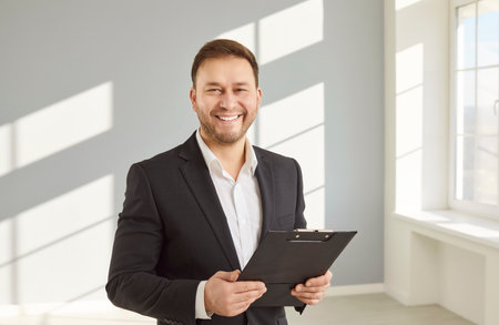 Portrait of young smiling attractive real estate agent man in suit with sale documents looking at camera standing in new empty apartments. Real estate market, relocation and moving concept.の写真素材