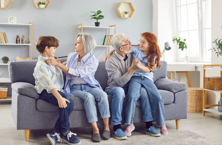 Happy cheerful senior grandparents sitting on sofa with grandchildren boy and girl in living room at home hugging and smiling enjoying leisure time together indoors. Generations and family concept.の写真素材