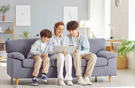 Mother and sons are spending leisure time together at home, sitting on sofa, looking through a...