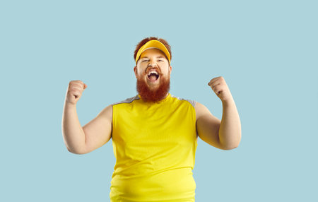 Cheerful crazy funny bearded fat man shouts loudly rejoicing in victory of his favorite sports team. Portrait of excited chubby man clenching his fists with joy isolated on light blue background.の写真素材