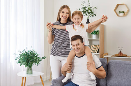Portrait of young happy family of three standing at home with their child girl looking at camera and smiling. Cute daughter sitting on fathers shoulders indoors. Love, care and happy family conceptの写真素材