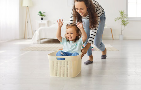 Happy mother and child playing together. Young mom pushing a plastic laundry basket with her cute little smiling cheerful kid daughter sitting inside. Family, leisure, fun conceptの写真素材