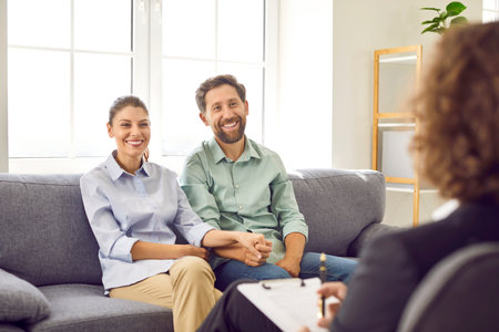 Cheerful family couple meeting with professional therapist or psychologist. Happy husband and wife having therapy session, sitting on couch, holding hands, talking about their relationship and smilingの写真素材
