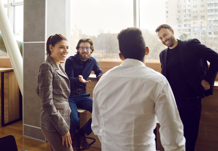Collegues talking in office during work break near window, smiling and friendly people. They discussing something looking at man in white shirt who sitting back to camera. Office lifestyle concept.の写真素材