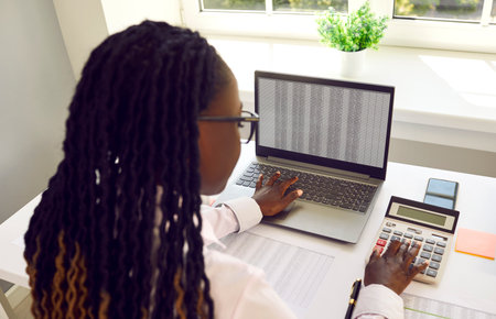 Young African American woman accountant working in the office, sitting at her desk, using a modern laptop computer and a calculator. Business, accounting concept. Back view over the shoulderの写真素材