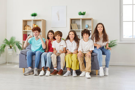 Happy excited preteen friends sitting on sofa, cheering their favorite football team. Cheerful classmates in colorful clothes spending leisure time together, watching TV and celebrating scored goal.の写真素材