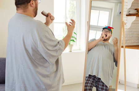 Funny man singing into brush before mirror. At home in pajamas and glasses he sings in cap using reflection as stage and brush as mic. Concept joyful playfulness and body confidence.の写真素材