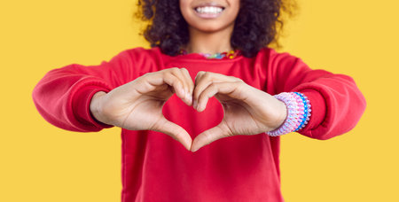 Crop closeup narrow banner show of happy African American girl show love sign. Smiling funny biracial kid demonstrate heart hand gesture share care and support. Yellow studio background.の写真素材