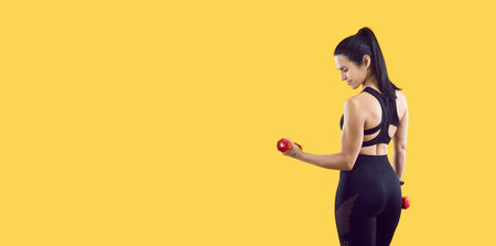 Healthy lifestyle. Smiling sporty woman with beautiful figure posing with dumbbells isolated on yellow background. Female fitness trainer in stylish and comfortable sportswear near copy space. Banner.の写真素材