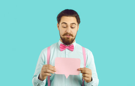 Portrait of a handsome man in a shirt and bow tie holding a rectangular pink mockup speech balloon. Young guy dressed up for a party showing a blank clean paper mock up message bubbleの写真素材