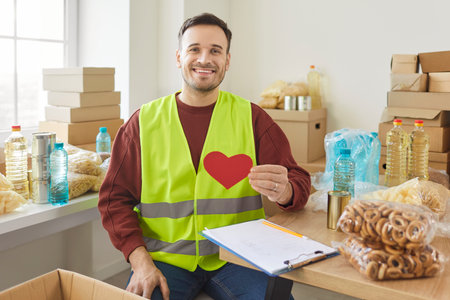 Portrait of smiling young man volunteer holding red heart in hands and looking at camera working at...