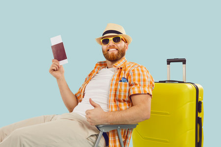 Happy man, male tourist summer beach wear holding passport, airline ticket, dreaming of flight. Planning holiday aircraft journey, travelling, sitting near large yellow suitcaseの写真素材