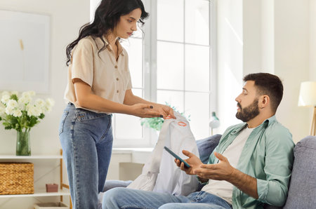 Couple arguing at home over smartphone. She confronts him with lipstick stain on shirt, jealousy rising with suspicion of infidelity. Fragile trust, family relationship conflict and betrayal.の写真素材