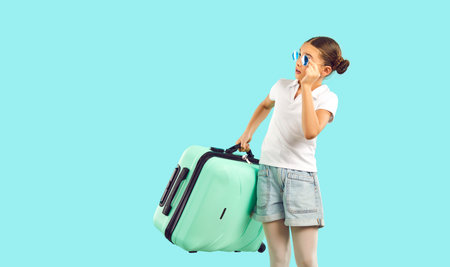 Child with suitcase going on summer vacation. Surprised tourist girl in shorts and T shirt standing isolated on bright turquoise background looking at copy space on left. Holiday, travel, kids conceptの写真素材
