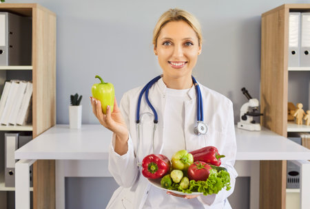 Nutritionist doctor promotes health with vegetables. In a clinic, she holds a green pepper and a bowl of produce, wearing stethoscope. Practical diet advice for preventative wellness.の写真素材