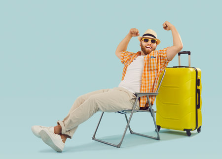 Vacation victory. Joyful funny man traveler sitting triumphantly with arms raised in victory near yellow suitcase. Man in casual summer clothes sits on folding chair isolated on light blue background.の写真素材