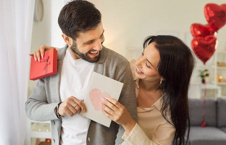 Portrait of a young pretty smiling woman holding greeting card and red present box and hugging her boyfriend. Happy couple in love. Care, congratulations, tenderness and Valentines day concept.の写真素材
