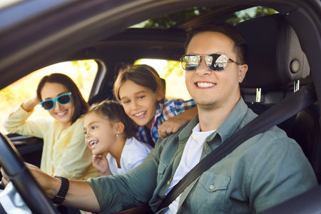 Happy family journey by car, road trip of parents and children. Tourists travel together, cheerful father wearing sunglasses and seatbelt sitting behind steering wheel, mother, son and daughter laughの写真素材