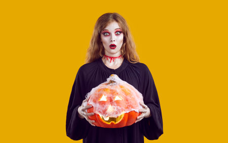 Blonde woman in witch costume with spooky makeup and fake bloody cut neck holding Halloween pumpkin covered with cobwebs and looking at camera with funny surprised face expressionの写真素材