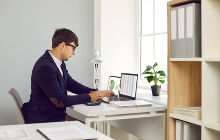 Businessman calculates business report, reviews company statistics, studies financial and marketing data. Man sits with calculator in front of laptops with graphs and spreadsheets open on screen.の写真素材