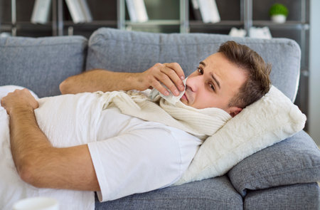 Sick man with fever, cold, virus, and tissue. He lies on a sofa, wiping nose with tissue, scarf on, resting to recover from cough, flu. Tea brings comfort. Concept: illness at home recovery.の写真素材