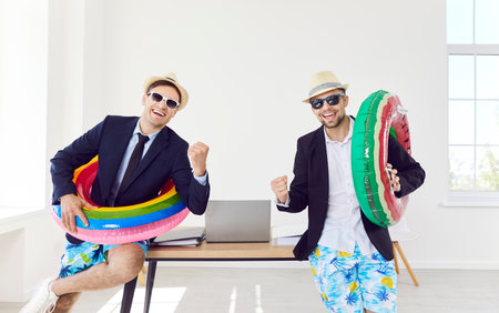 Funny business male workers in sunglasses and beach hats standing at desk in office with rubber ring going on holiday trip and ready for summer vacation. Travel, journey and travelling concept.の写真素材
