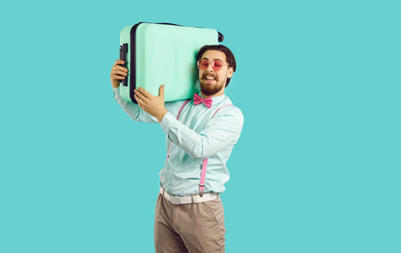 Handsome stylish young man holding suitcase on his shoulders on light blue background. Caucasian guy with beard and mustache wearing shirt, suspenders, bow tie and glasses looks at camera. Isolated.の写真素材