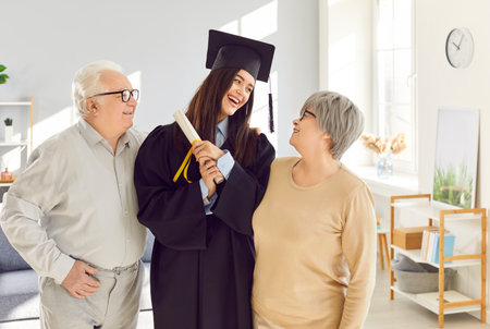 Young happy girl wearing university graduate gown holding diploma in hands standing with her senior old parents proud of her in living room at home. Graduation and education concept.の写真素材