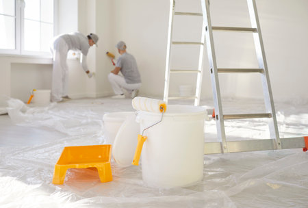 Step ladder, bucket with paint roller standing in empty light room in new house with male painters team from repair service painting a wall in background. Renovation process concept.の写真素材