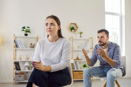 Quarrel and fight of couple at home, angry husband arguing with gestures while wife ignoring in relationship crisis. Argument and tension from man to sad woman sitting in living room with frustrationの写真素材