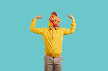 Man in funny chicken disguise pretends to be strong and have superpower. Overconfident young guy wearing strange ridiculous bizarre rooster mask flexing his weak arms isolated on turquoise backgroundの写真素材