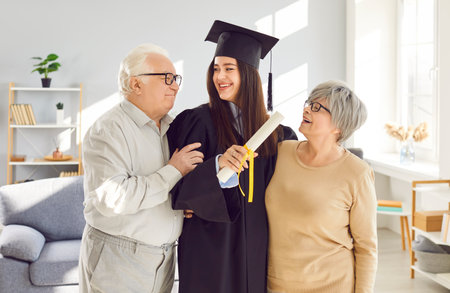Happy and proud elderly parents are congratulating adult graduate daughter in mortarboard and gown, holding diploma at home. The daughter is happy and proud, celebrating graduation with parents.の写真素材