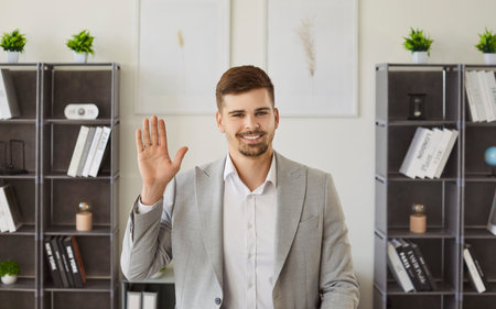 A young man wearing formal clothes looking at camera waving hand having video call standing on workplace in office. Confident man chatting online or having video conference call. remote job concept.の写真素材