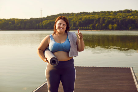 Young happy smiling funny woman in sportswear showing thumb up sign celebrating success in workout standing on pier outdoors in the park. Healthy lifestyle, sport and fitness concept.の写真素材