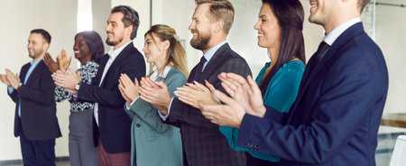 Diverse happy smiling young business people in suits standing in a row on conference and listening to colleague or leadership. Company employees applauding on meeting in office.の写真素材