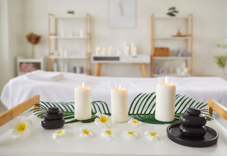 Serene spa setting with burning candles, black stones and white flowers arranged on table. Modern interior of massage or cosmetology room with relaxing decor and massage table on background.の写真素材