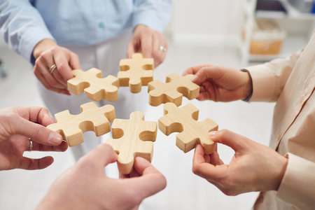 Close up shot of a hands of a business people team holding wooden puzzle pieces. Teamwork, communication, and collaboration within a company or partnership, as they work together to solve a goal.の写真素材