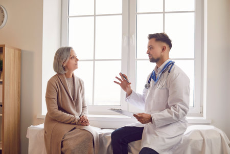 Portrait of elderly gray-haired woman in medical office listening to man doctor holding report file with appointment during medical exam in clinic. Health care and medicine concept.の写真素材