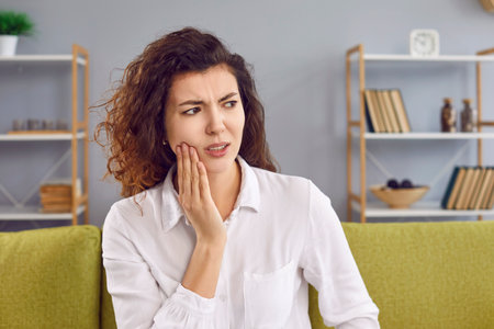 Portrait of a young attractive upset woman sitting on the sofa in the living room at home suffering from toothache. Curly redhead sad girl in a white shirt holding her cheek in dental pain.の写真素材