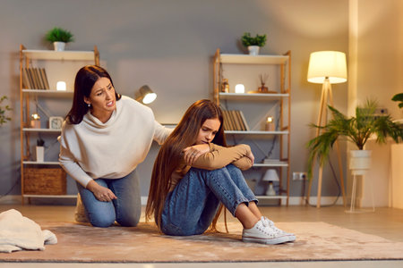 Caucasian mother is angry and scolding crying, frustrated daughter, sitting on floor at home living room in the evening, engaging in a conflict as part of parenting and resolving family issues.の写真素材