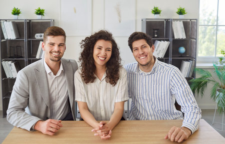 Three cheerful smiling young coworkers sitting at desk in office and looking at camera. Happy male...