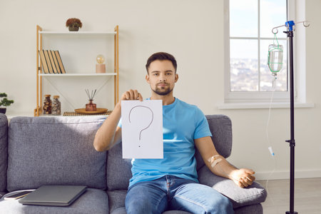 Patient doubting effectiveness of treatment therapy. Young man sitting on couch at home, getting intravenous IV line infusion and showing sheet of paper with question mark. Medical uncertainty conceptの写真素材
