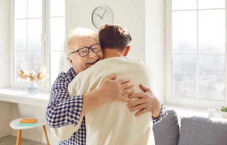 Happy smiling gray-haired old senior parent hugging his adult son in living room. Young man embracing his elderly father visiting him at home. Love, family and fathers day concept.の写真素材