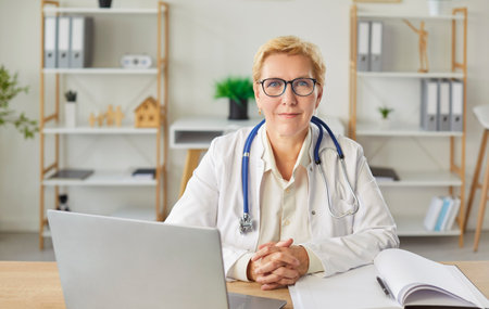 Mature female doctor smiling at desk, medical consultant, female medical nurse or practitioner, clinic manager analyzing data, keeping patient information, health center assistant, client adviceの写真素材
