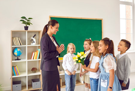 Teacher day. Children singing song congratulating happy teacher in school. Elementary school students greeting their teacher in classroom with bouquet of flowers. Back to schoolの写真素材